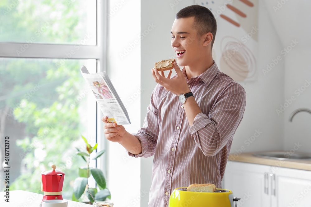 Young man eating toast with tasty nut butter and reading newspaper in kitchen