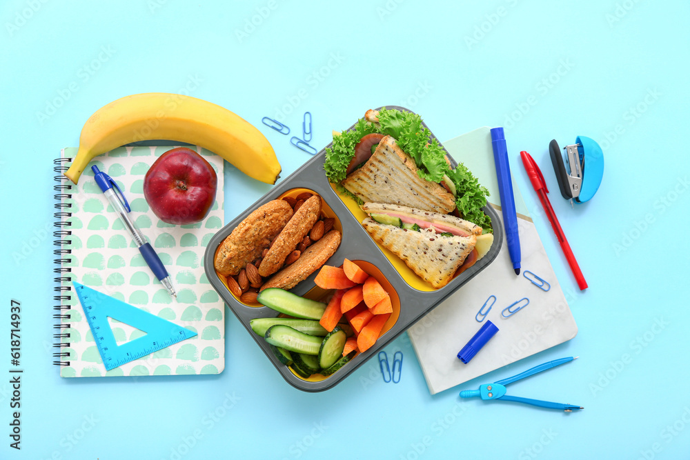 Stationery and lunch box with tasty food on blue background