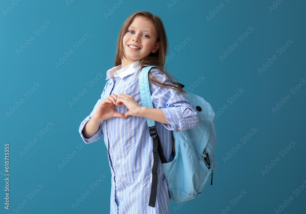 Little schoolgirl with backpack making heart gesture on blue background