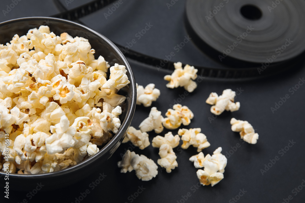 Bowl with tasty popcorn and film reel on black background