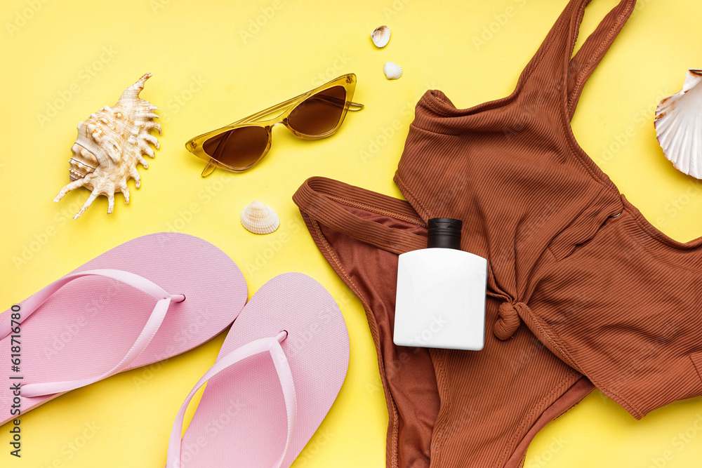 Pink flip-flops with bottle of sunscreen cream, swimsuit and sunglasses on yellow background