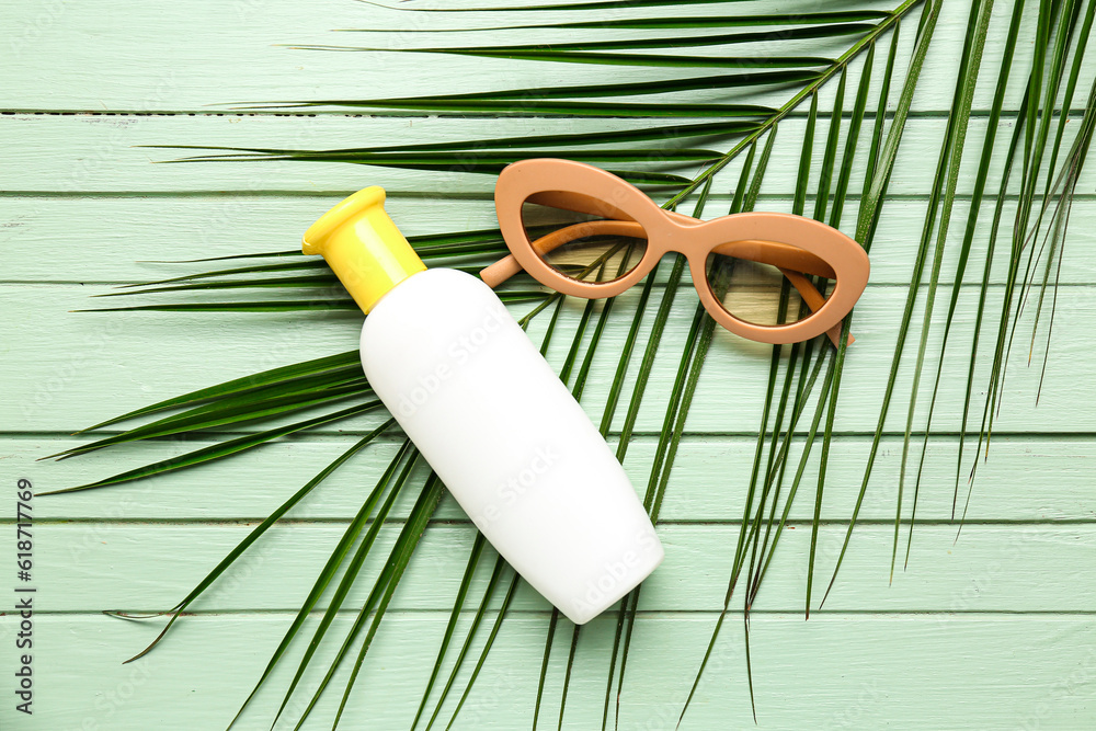 Sunglasses with bottle of sunscreen cream and palm leaf on green wooden background