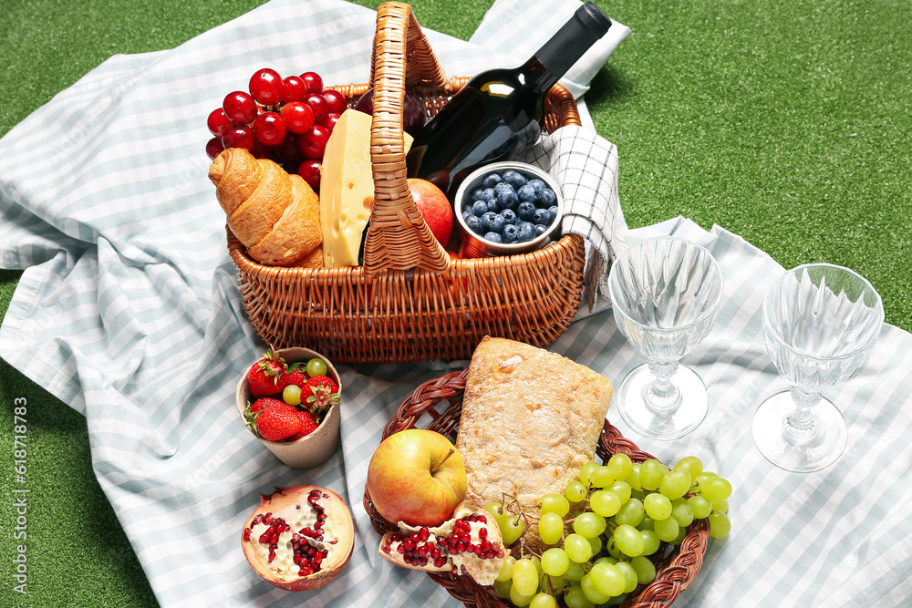 Wicker basket with tasty food for picnic, wine and glasses on green background