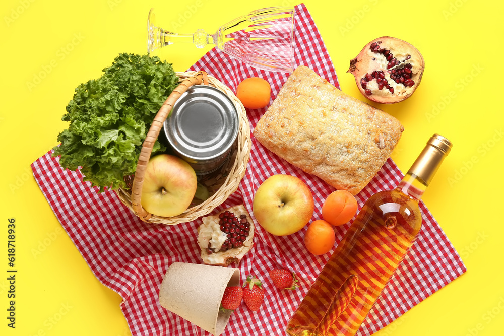 Wicker basket with tasty food for picnic, wine and glass on yellow background