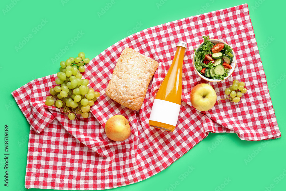 Bowl with salad, bottle of juice, bread and fruits for picnic on green background