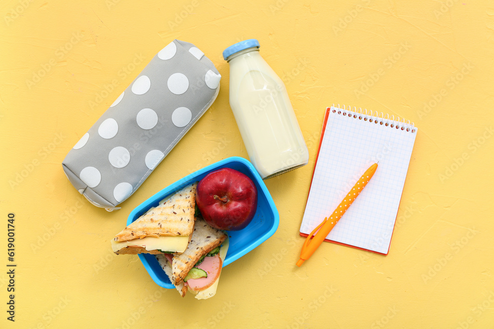 Stationery, drink and lunch box with tasty food on yellow background