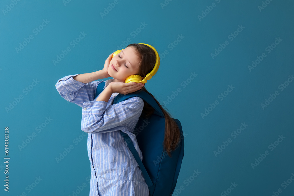 Little schoolgirl in headphones with backpack on blue background