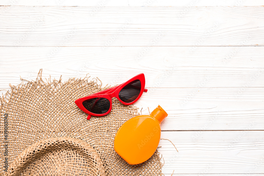 Wicker hat with sunglasses and bottle of sunscreen cream on white wooden background