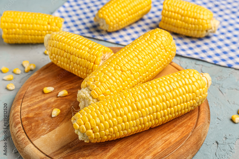 Wooden board with fresh corn cobs and seeds on blue background