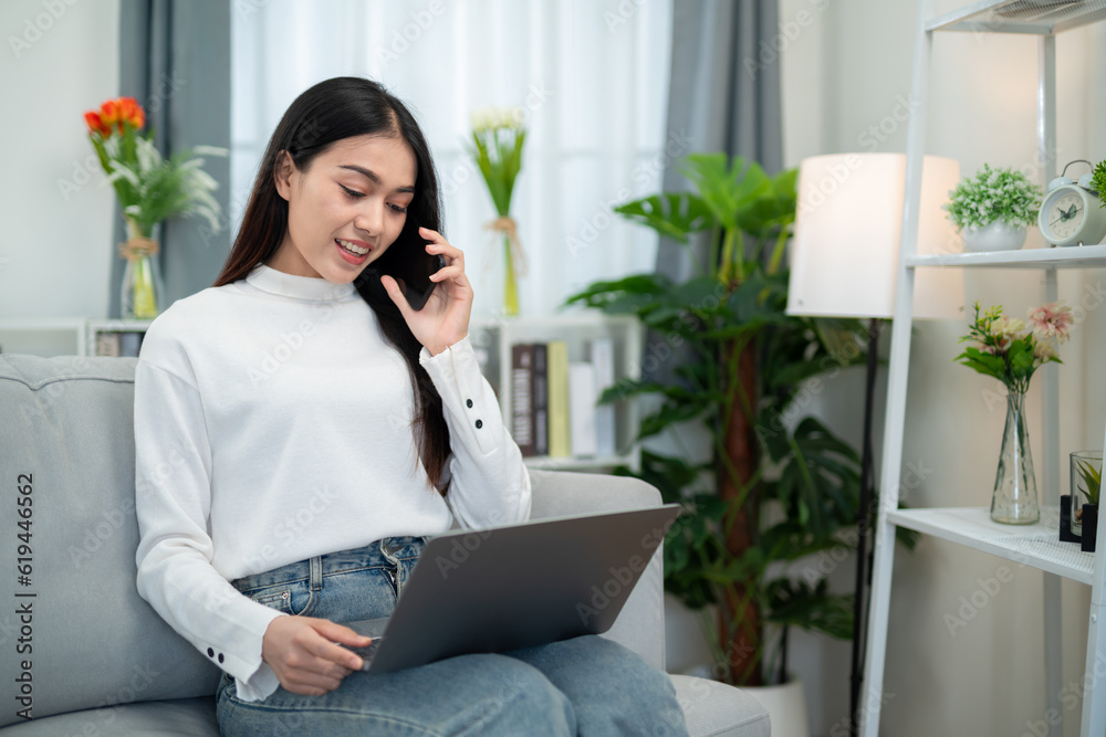 Asian woman talking on the phone while checking online data with laptop on sofa in living room. Work