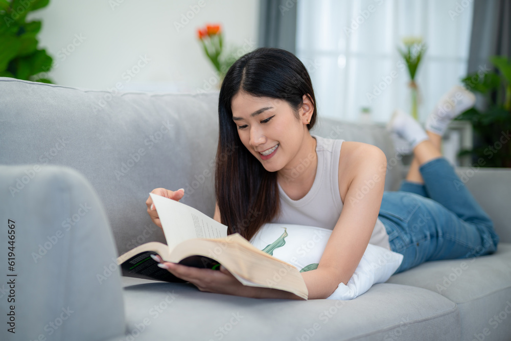 Asian woman lying and reading a book on the sofa in the living room in the morning.