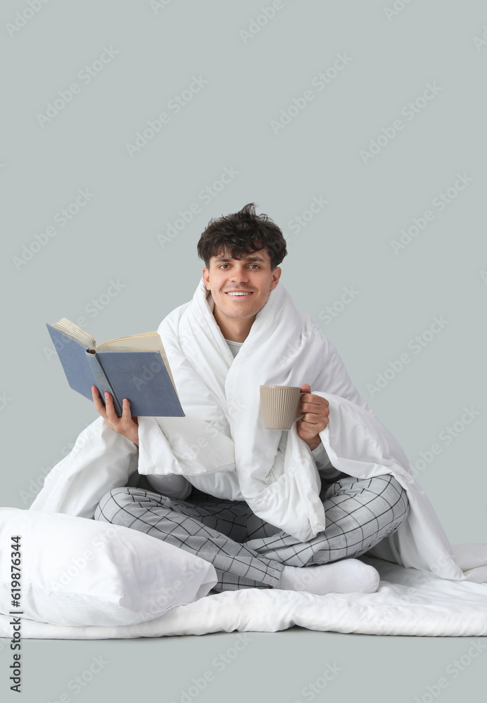 Handsome happy man with cup of coffee, soft blanket and pillow reading book on grey background