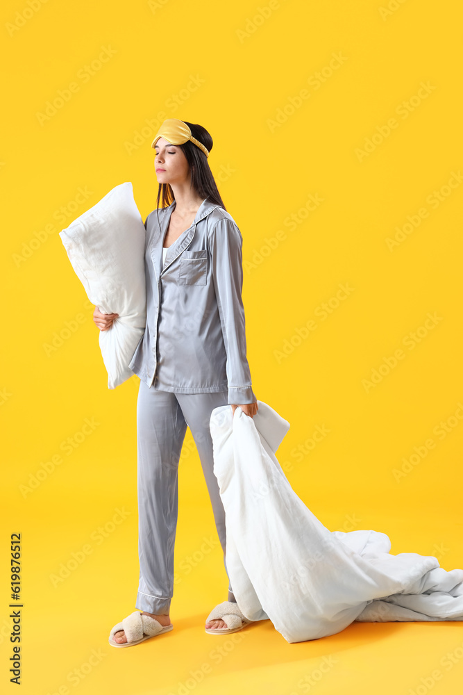 Young woman in pajamas with pillow and blanket on yellow background