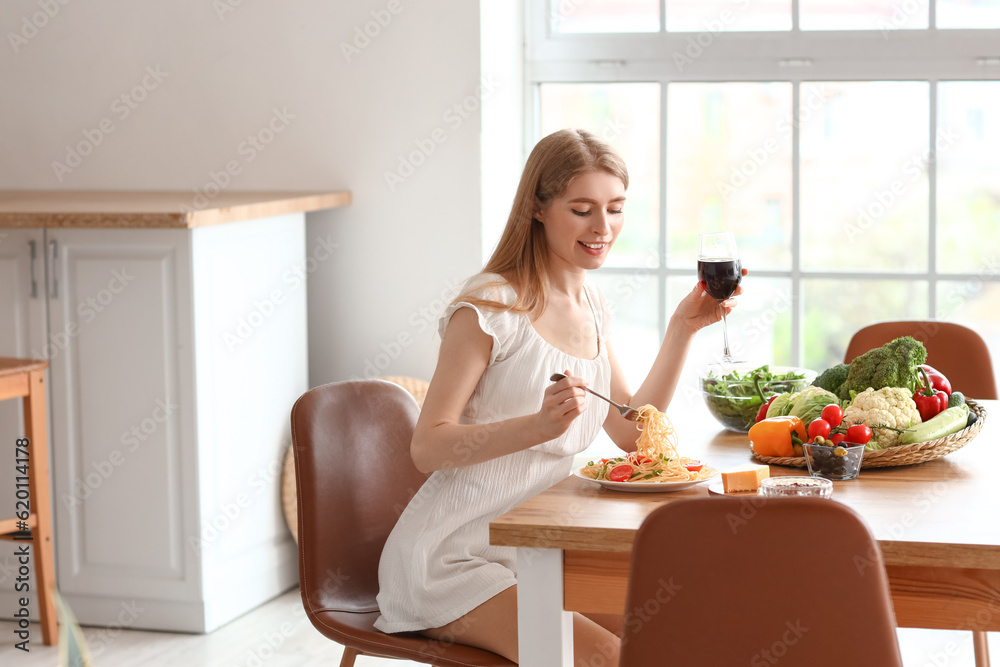 Young woman with glass of wine eating tasty pasta in kitchen