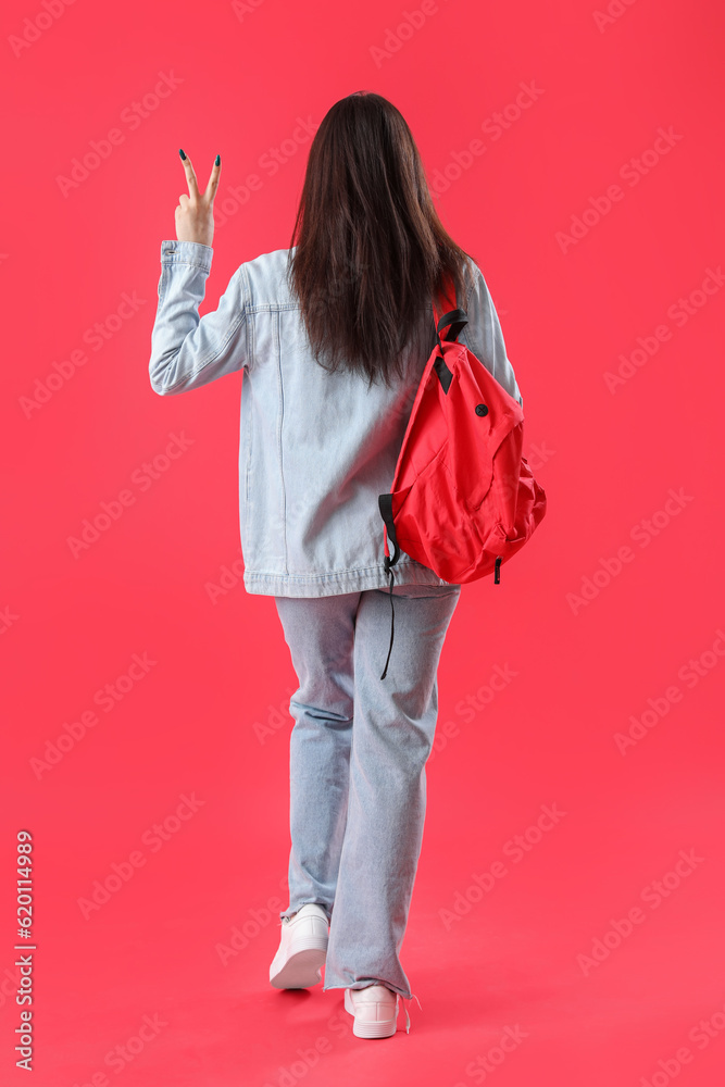 Female student with backpack showing victory gesture on red background, back view