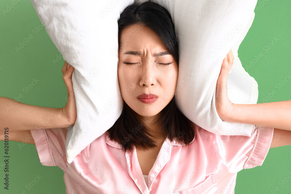 Displeased Asian woman in pajamas with pillow on green background, closeup