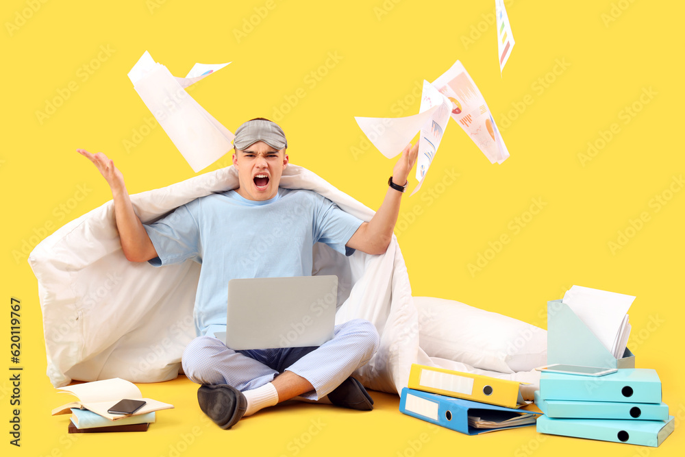 Stressed young man in pajamas, with documents and laptop working on yellow background