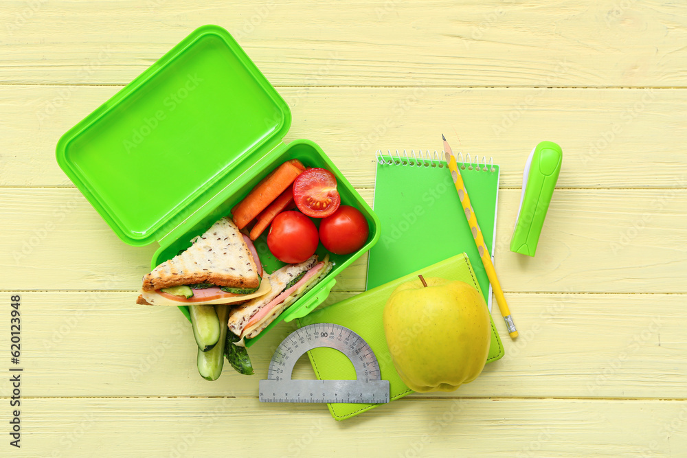 Stationery and lunch box with tasty food on yellow wooden background