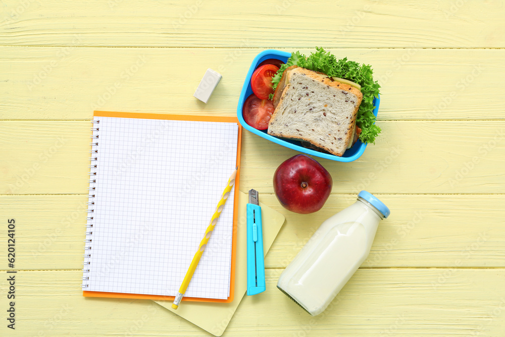 Stationery, drink and lunch box with tasty food on yellow wooden background