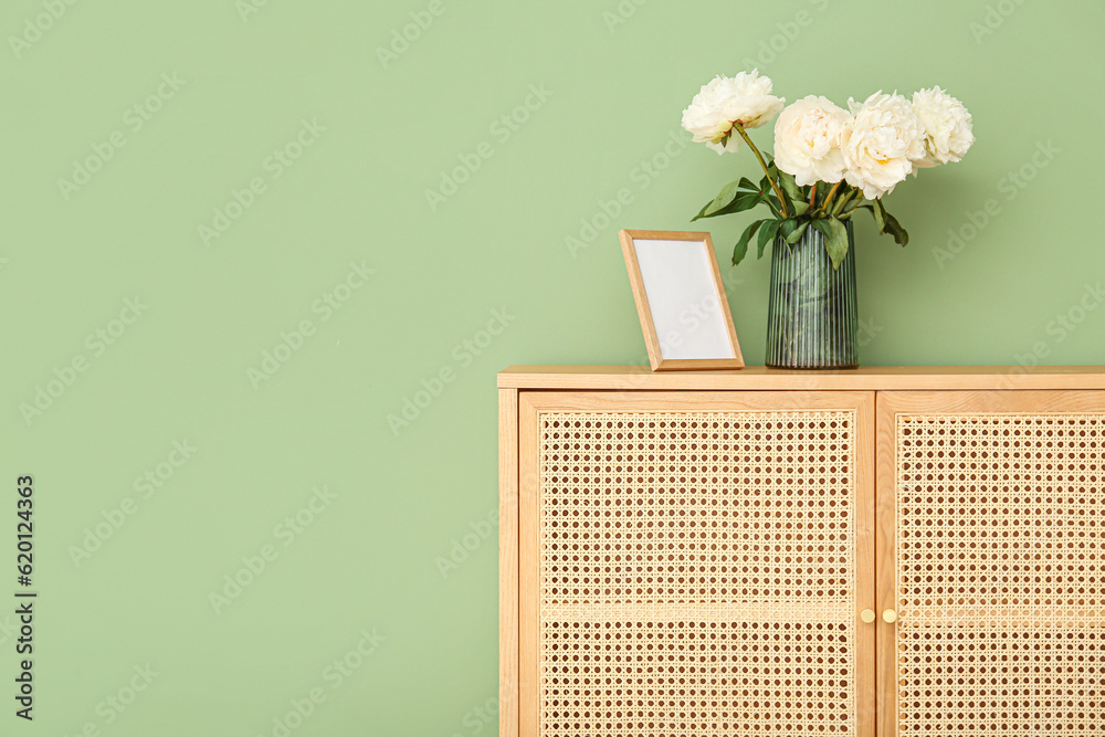 Vase of white peonies and photo frame on dresser near green wall