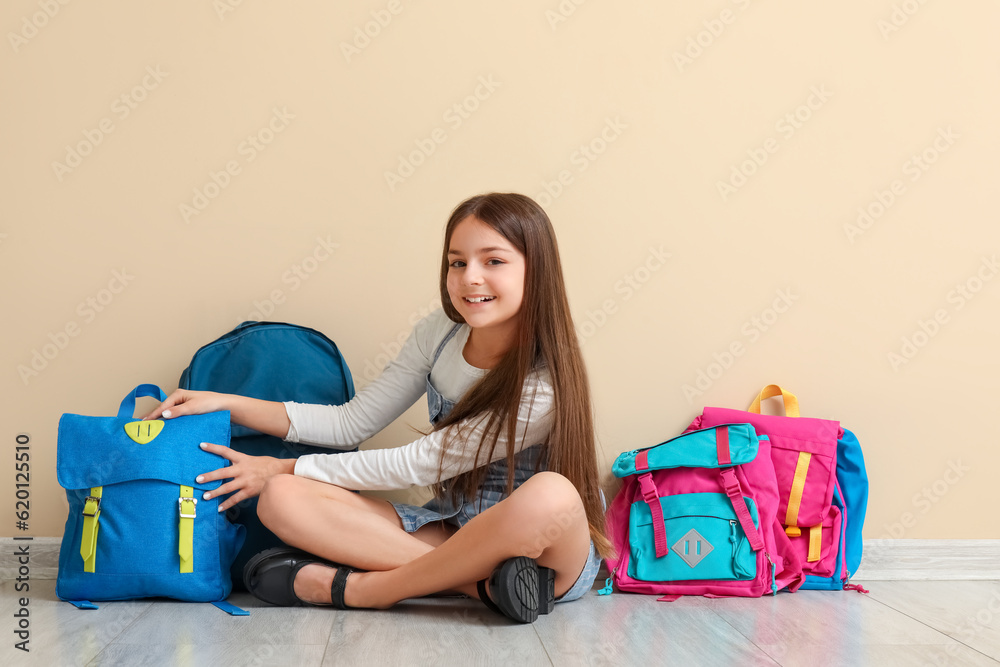 Little schoolgirl with backpacks sitting near beige wall