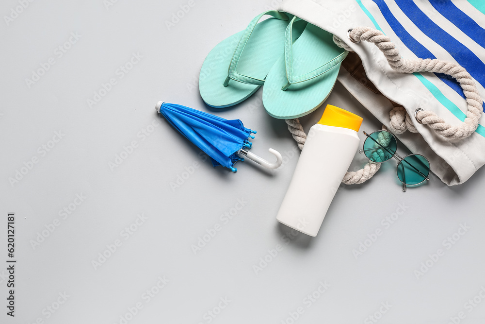 Bag with sunglasses, flip flops, mini umbrella and bottle of sunscreen cream on grey background