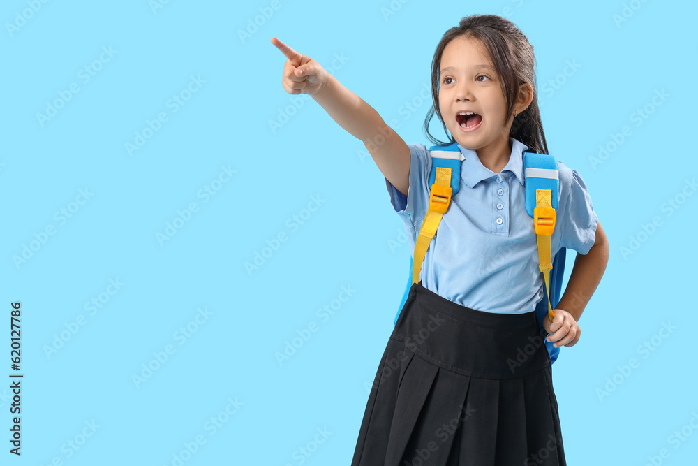 Little schoolgirl with backpack pointing at something on light blue background