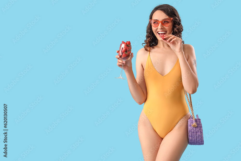 Young woman in swimsuit with glass of strawberries on blue background