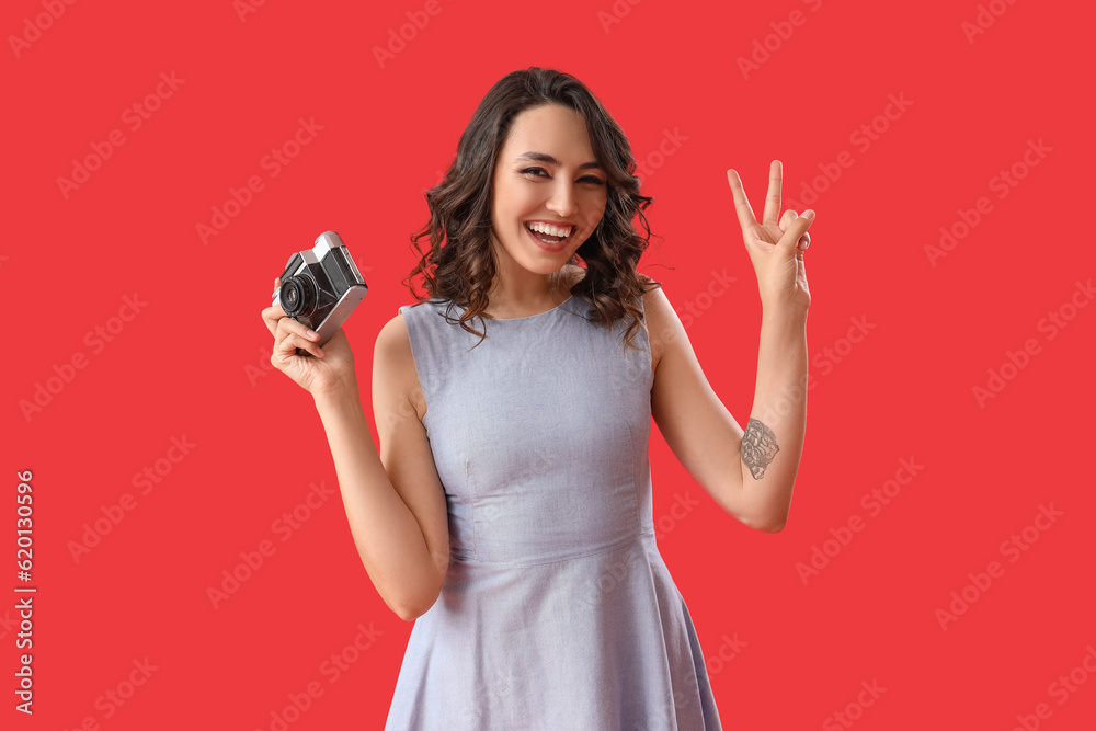 Young woman with photo camera showing victory gesture on red background