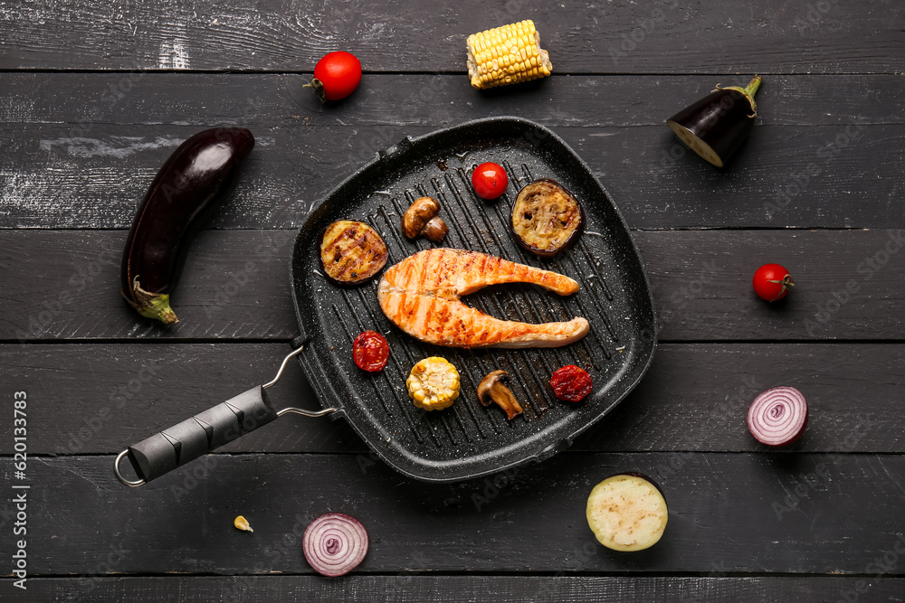 Frying pan with tasty grilled salmon steak and vegetables on black wooden background