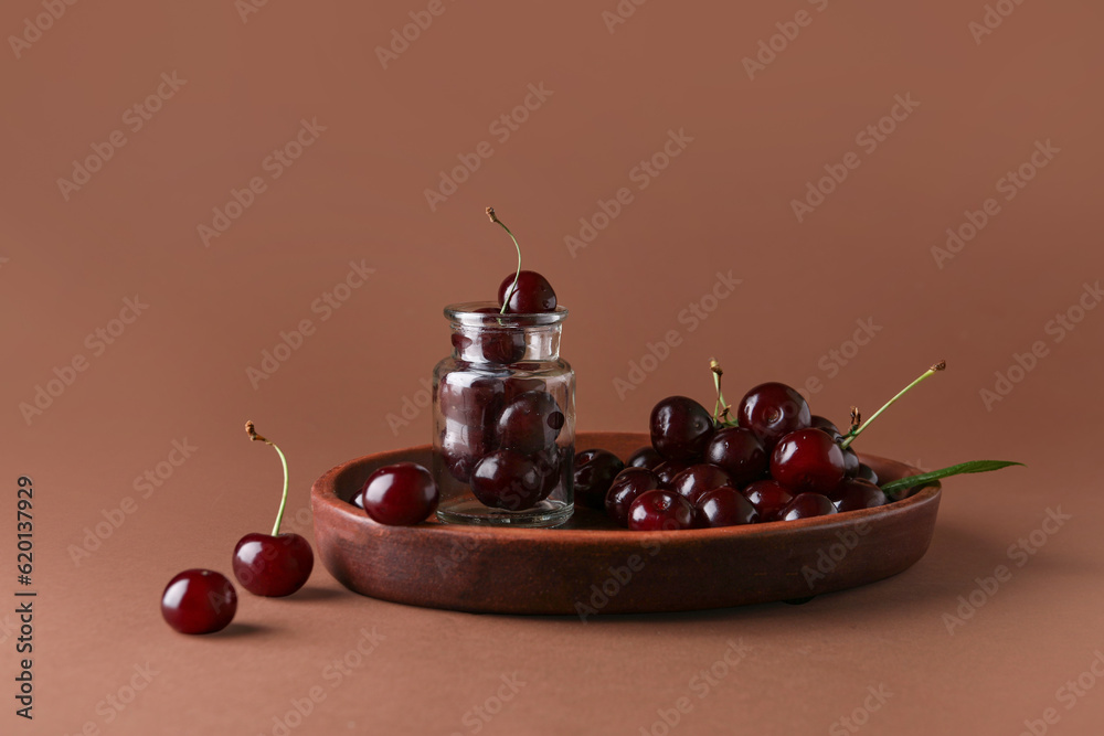 Jar and plate with sweet cherries on brown background