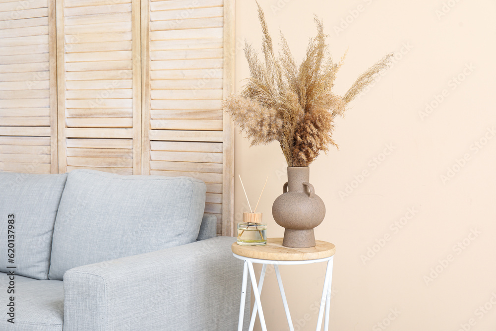 Table with aroma diffuser, pampas grass and sofa near beige wall in room