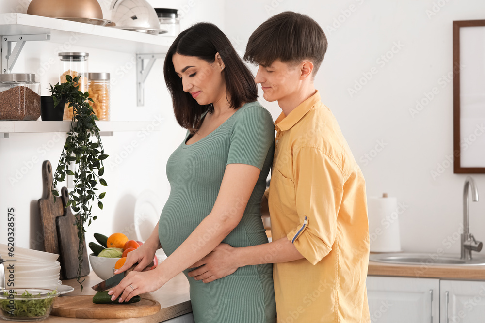 Young pregnant woman with her husband cutting cucumber in kitchen