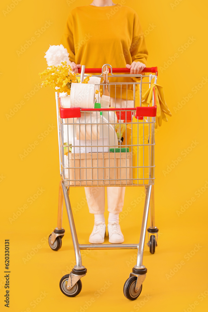 Woman with shopping cart full of cleaning supplies with flowers on yellow background