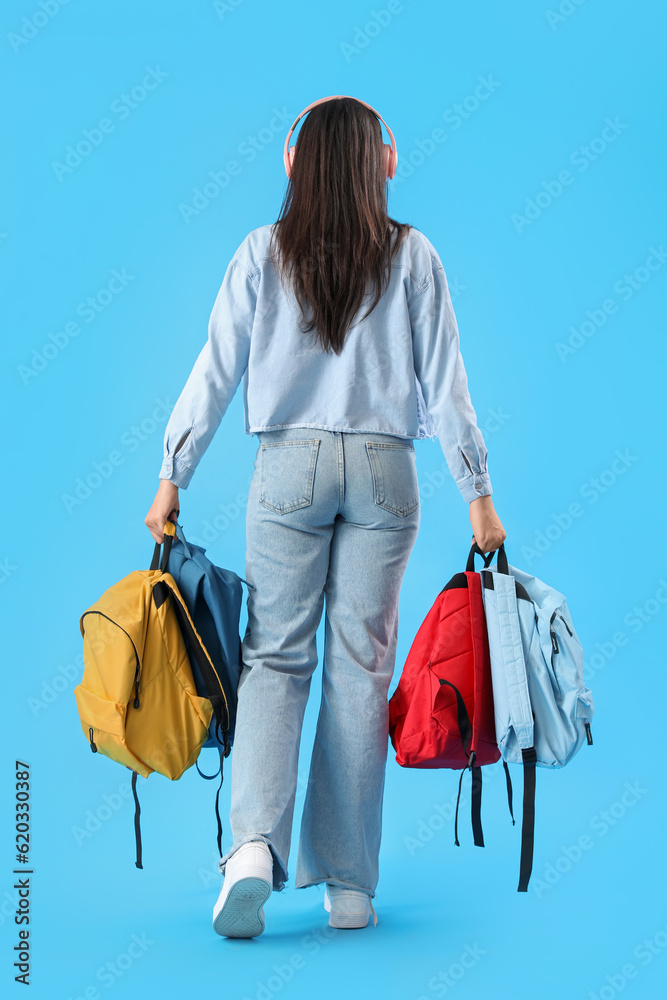 Female student in headphones with backpacks on blue background, back view