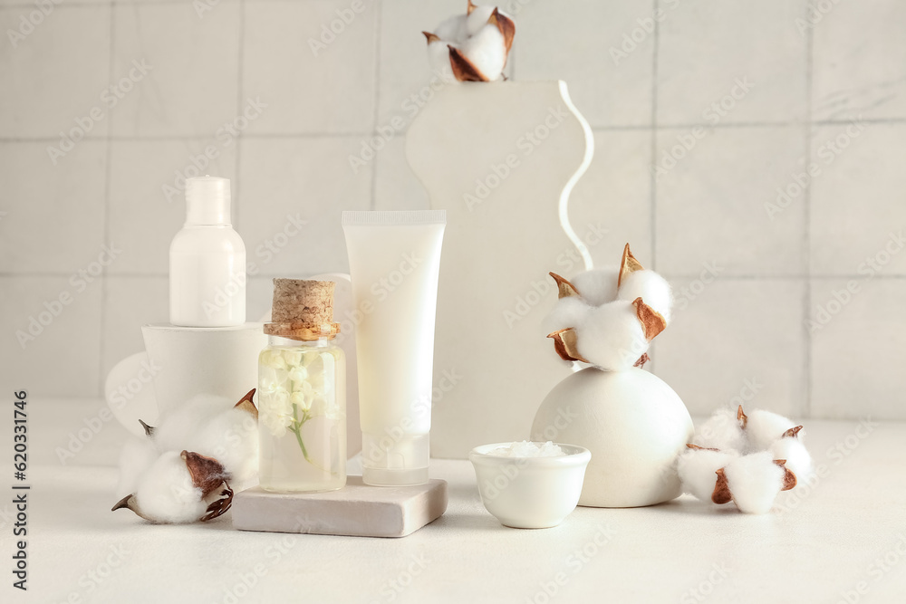 Composition with cosmetic products, plaster stands and cotton flowers on table against light tile ba