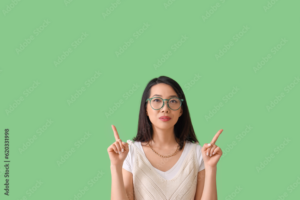 Beautiful Asian woman in stylish eyeglasses pointing at something on green background