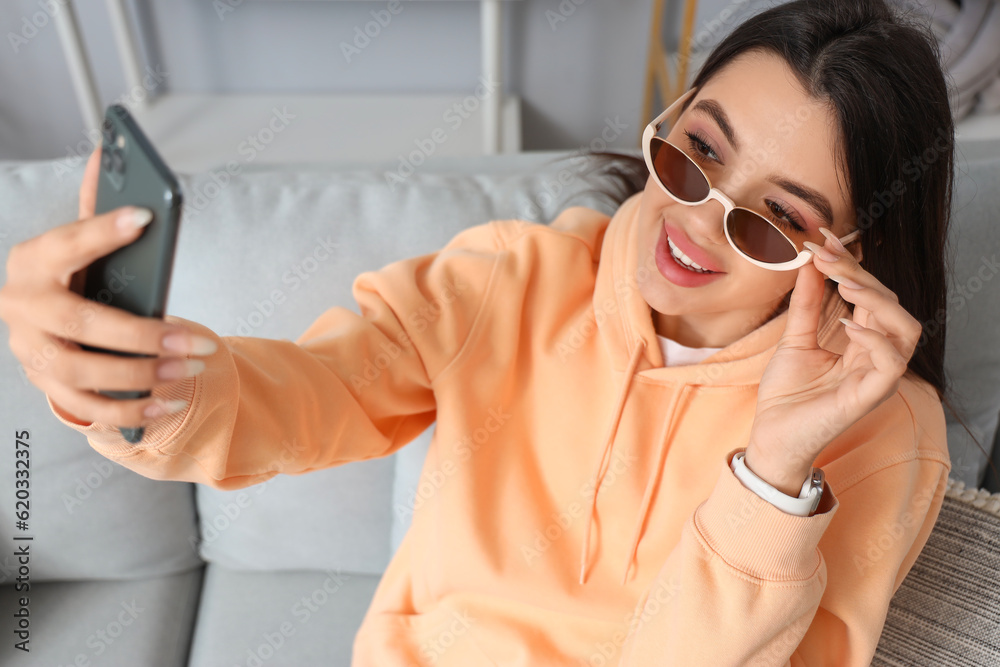 Young woman wearing stylish sunglasses while taking selfie at home