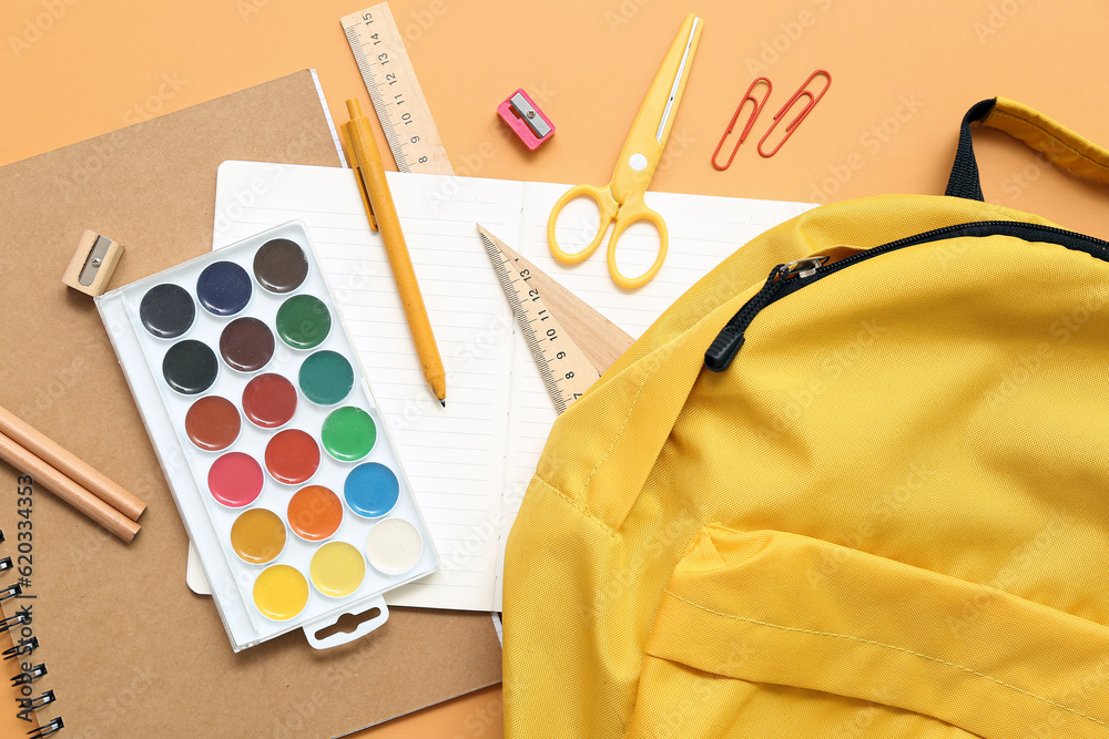 School backpack with paints and stationery on orange background