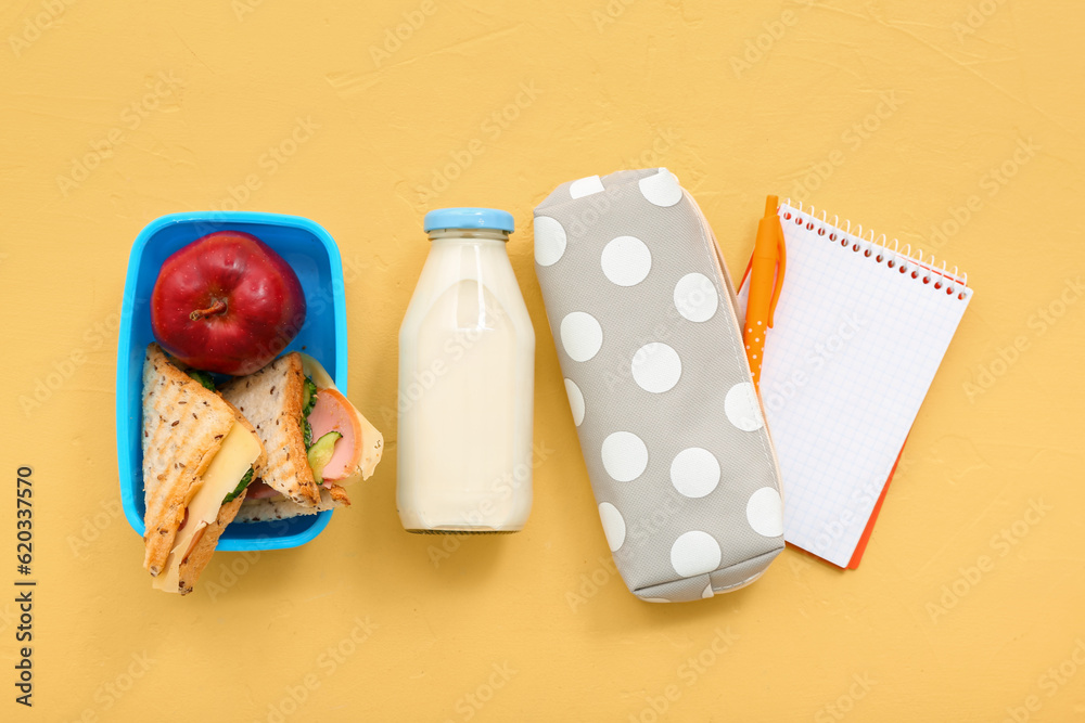 Stationery, drink and lunch box with tasty food on yellow background