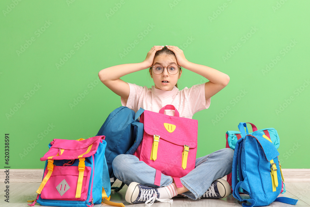 Shocked little girl with backpacks sitting near green wall