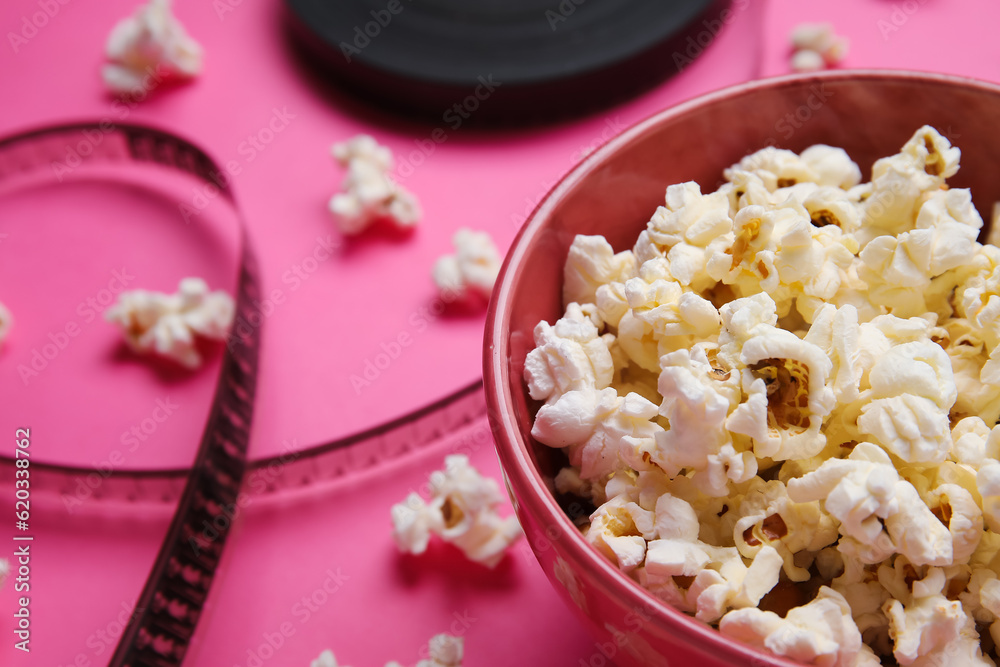 Bowl with tasty popcorn and film reel on pink background