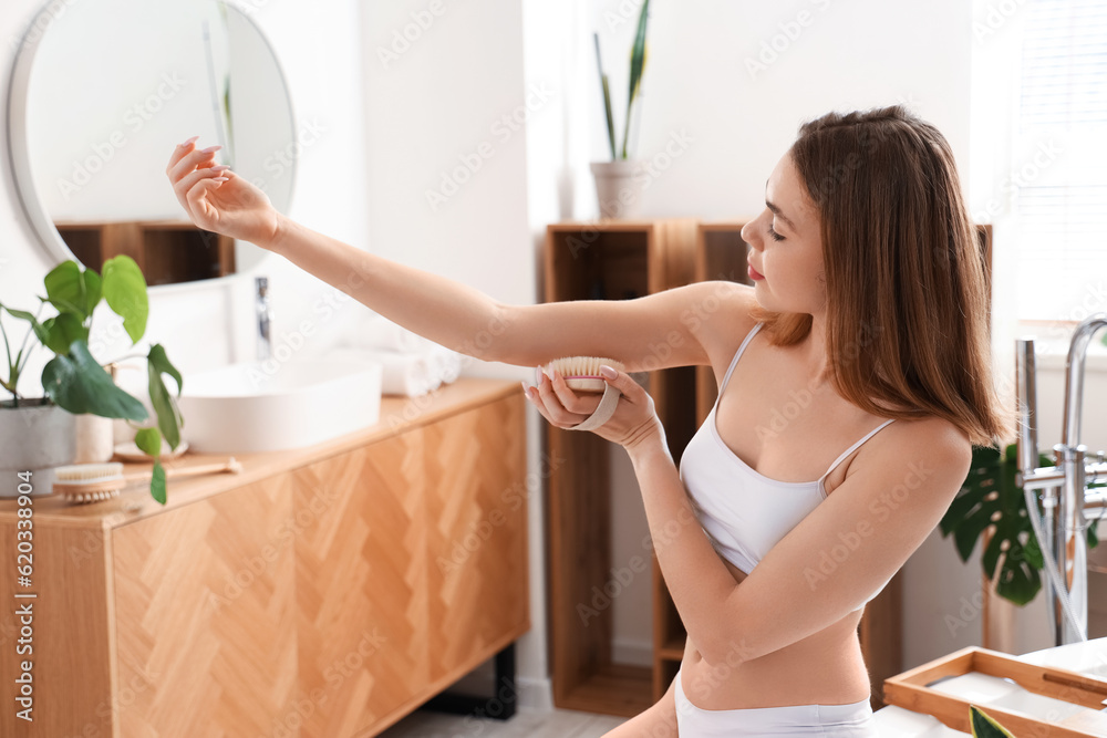 Young woman massaging her arm with anti-cellulite brush in bathroom