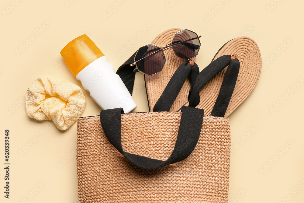 Wicker bag with sunglasses, flip flops and bottle of sunscreen cream on pale yellow background