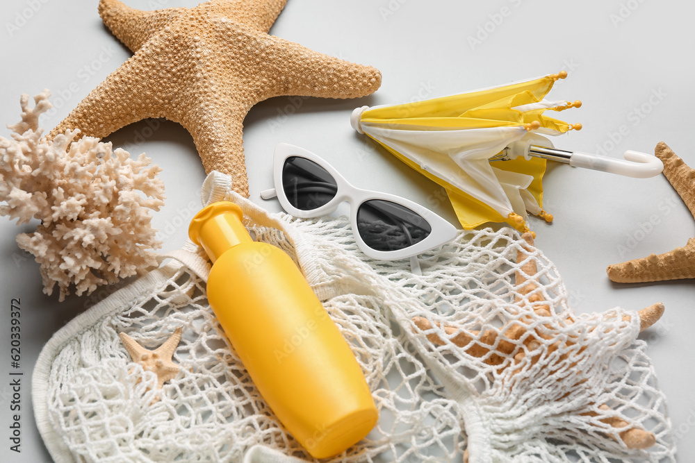 String bag with sunglasses, mini umbrella, coral and bottle of sunscreen cream on grey background