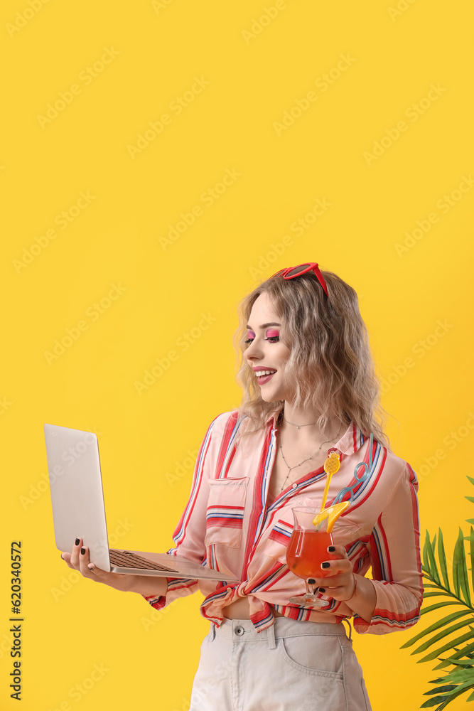 Young woman with laptop and summer cocktail on yellow background