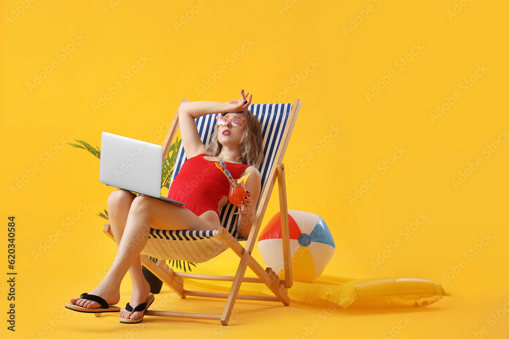 Upset young woman with cocktail and laptop in deck chair on yellow background