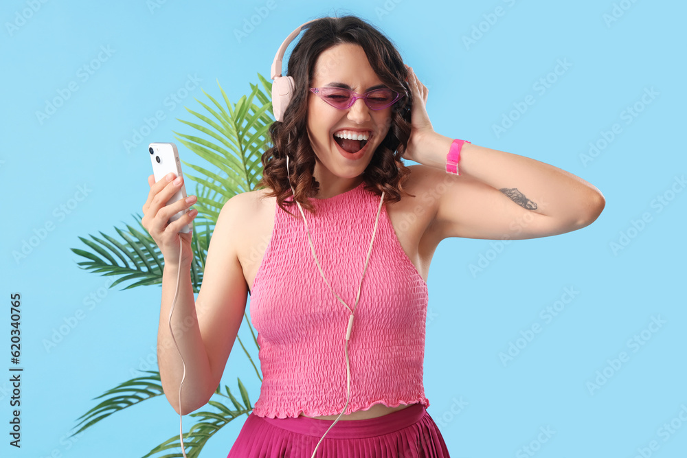 Happy young woman in headphones with mobile phone and palm leaves on blue background