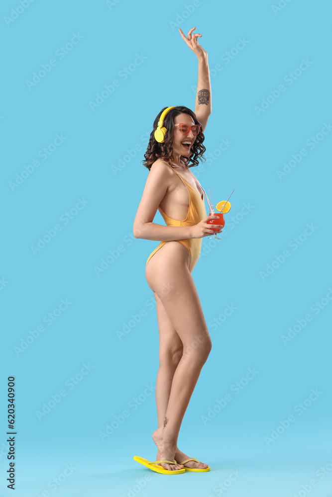 Young woman in swimsuit with headphones and cocktail dancing on blue background