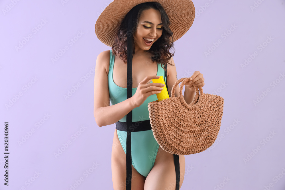 Young woman with sunscreen cream in beach bag on lilac background